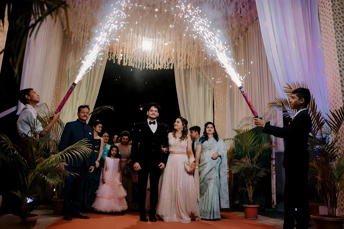 Bride and groom walking under sparklers at wedding entrance with family, festive drapes and palm decor