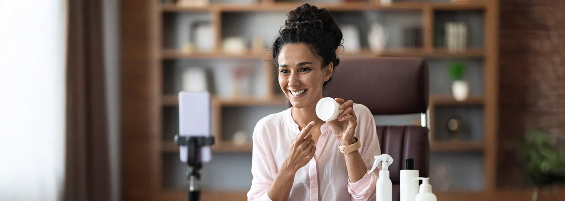Woman smiling while holding skincare jar during beauty tutorial, with product bottles and phone stand visible