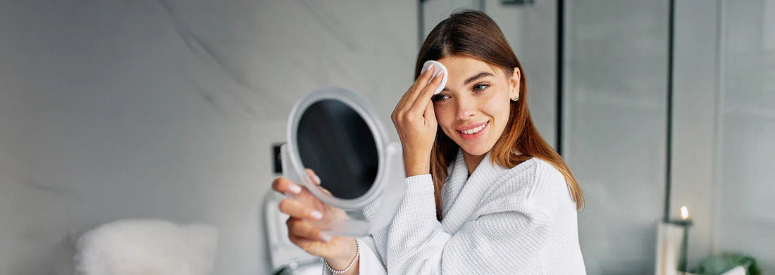 Woman using cotton pad to cleanse face in bathroom, holding round mirror, skincare routine