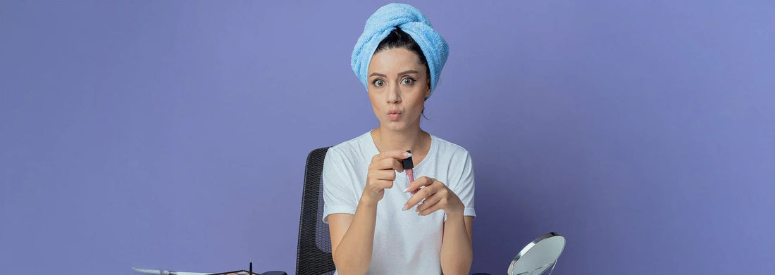 Woman applying lip gloss with blue towel on head, sitting at vanity with mirror against purple background