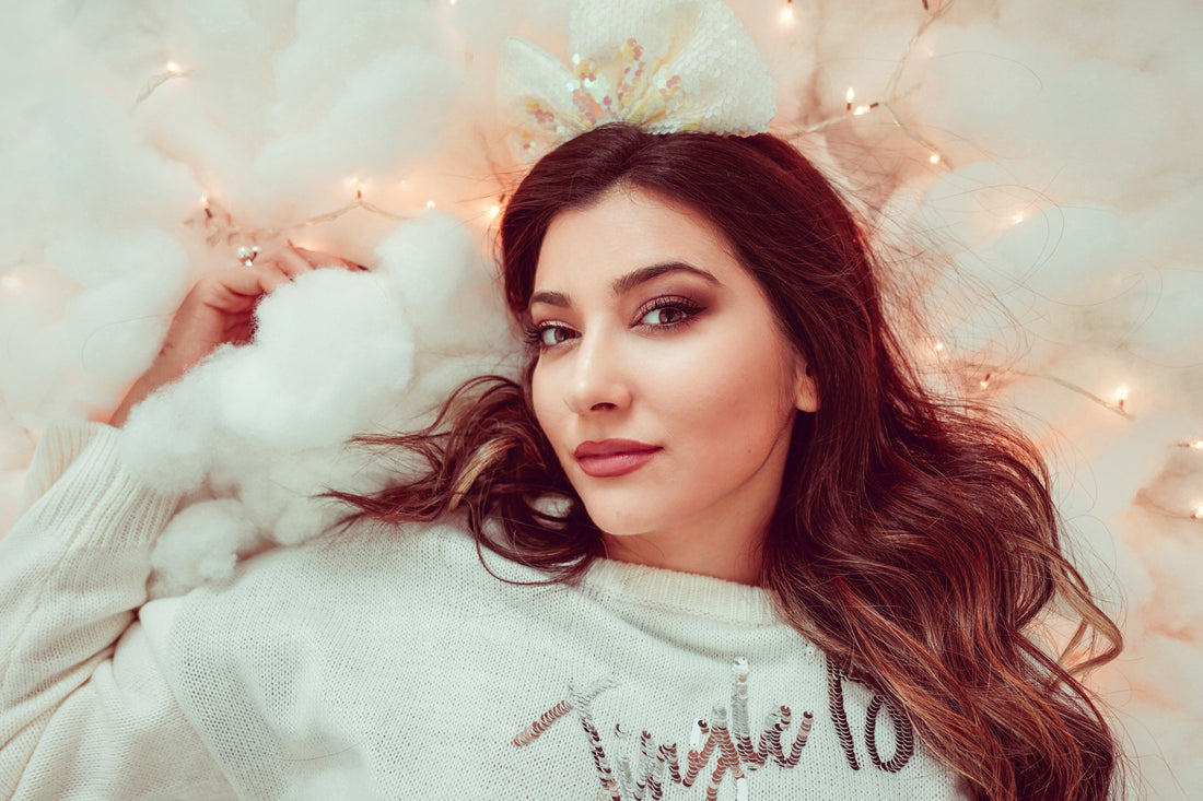 Woman with smoky eye makeup and nude lips wearing white sequin sweater, festive fairy lights background