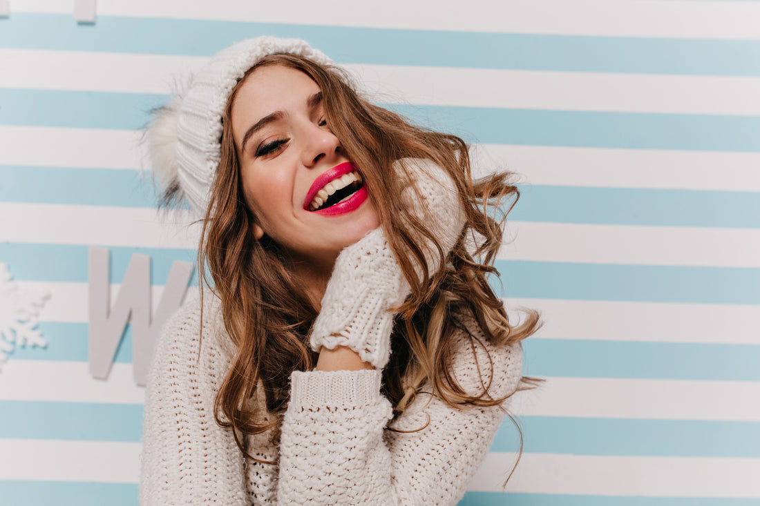 Woman laughing with bold pink lipstick, wearing white knit hat and sweater against blue-white striped wall